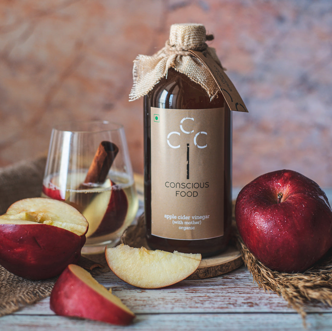 This image showcases a bottle of 'Conscious Food' organic apple cider vinegar with apples and a glass; rustic still life highlighting 'with mother' ACV.